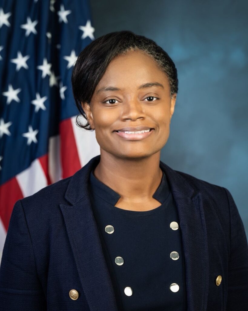 Professional headshot of Dr. Lutheria Peters, smiling, wearing a navy blazer with gold buttons, with an American flag in the background.