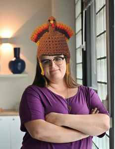 Woman with folded arms wearing a brown crocheted turkey hat with a red, orange, and brown tail fan and yellow earflaps.